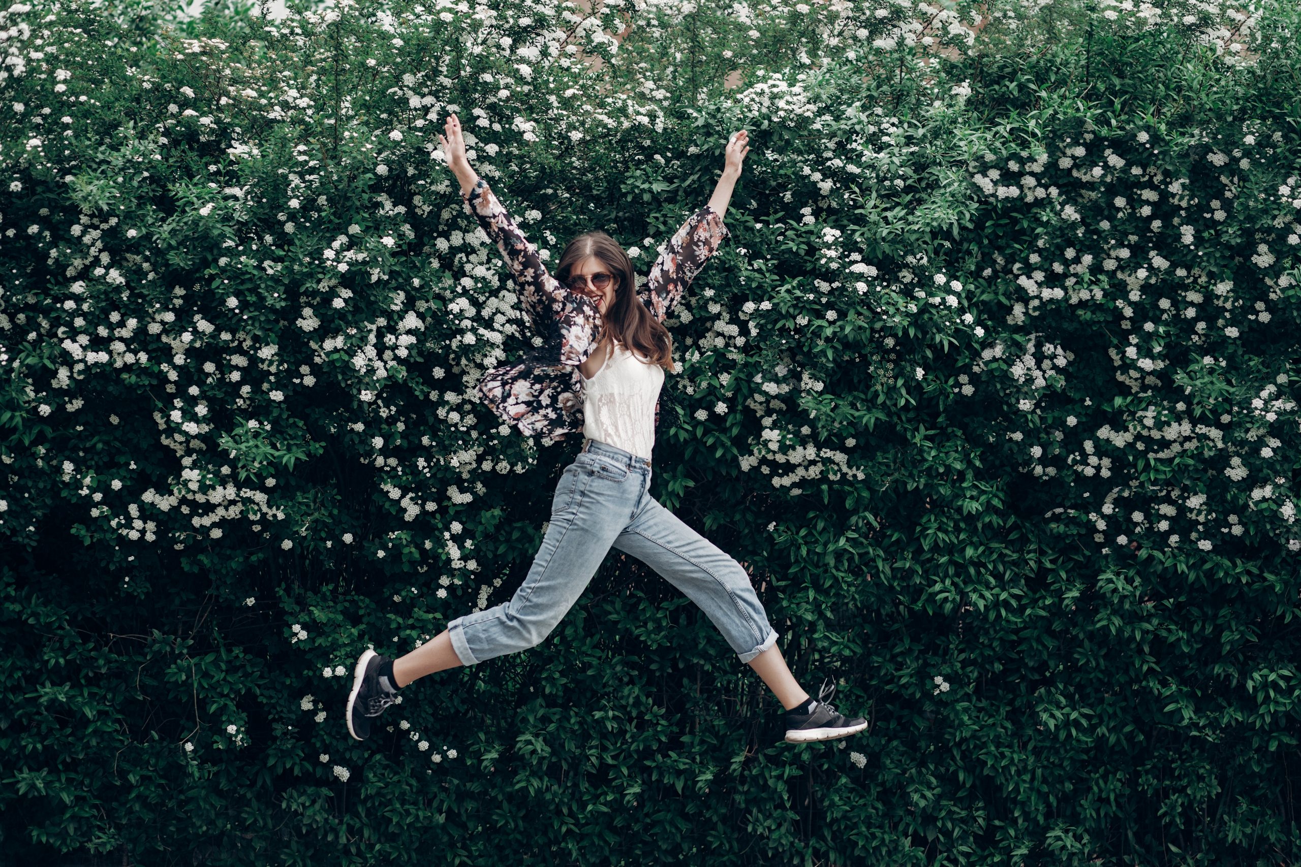 happy hipster woman in sunglasses having fun on background of blooming bush with white flowers of spirea. boho girl jumping and smiling in modern clothes, emotional moment. space for text happy hipster woman in sunglasses having fun on background of blooming bush with white flowers of spirea. boho girl jumping and smiling in modern clothes, emotional moment. space for text