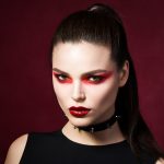 Fesche Gothic Frau mit heller haut und roten Lippen, die ein schwarzes Lederhalsbald mit Nieten trägt. Rote Smokey Eyes. Vampir oder Halloween Make-up.