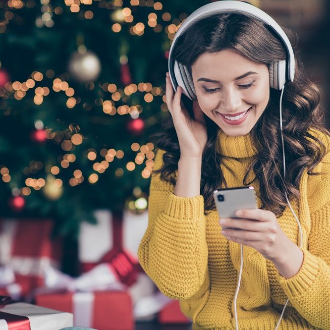 Photo of charming beautiful wavy curly haired girl listening to music, wearing yellow pullover staring at telephone screen