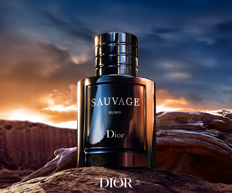 Eine Flasche Dior Sauvage Elixir Parfüm liegt auf einer felsigen Oberfläche, vor einem dramatischen Sonnenuntergangshimmel.
