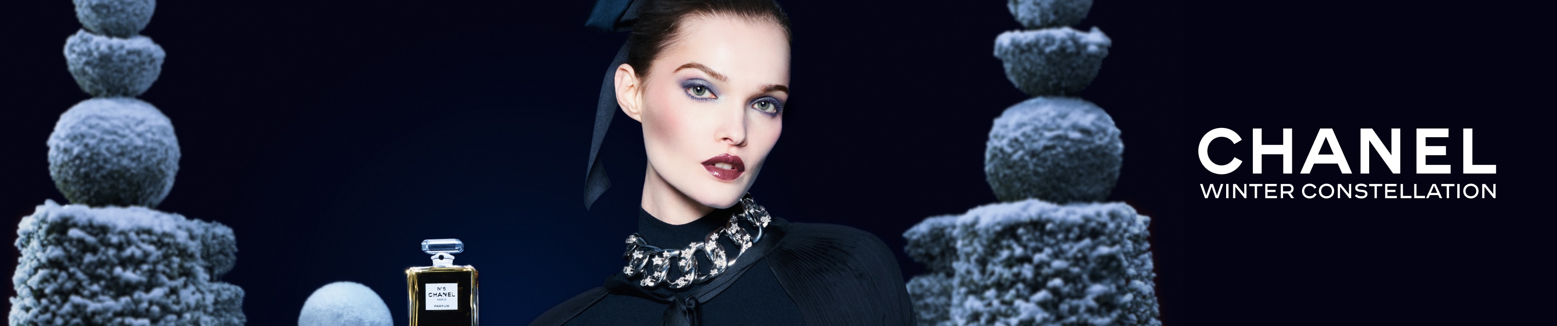 Chanels Winter Constellation-Kampagne mit einem Modell, das eine Statement-Halskette und dunkle Kleidung trägt, neben einer Flasche Chanel No. 5 Parfum. Im Hintergrund sind schneebedeckte Steinskulpturen vor einem dunkelblauen Hintergrund zu sehen