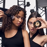 Zwei Frauen posieren vor Metallträgerstrukturen. Eine hält einen Make-up-Pinsel an ihr Gesicht, die andere hält den Lancôme Teint Idole Ultra Wear Bronzepuder vor ihr Auge.