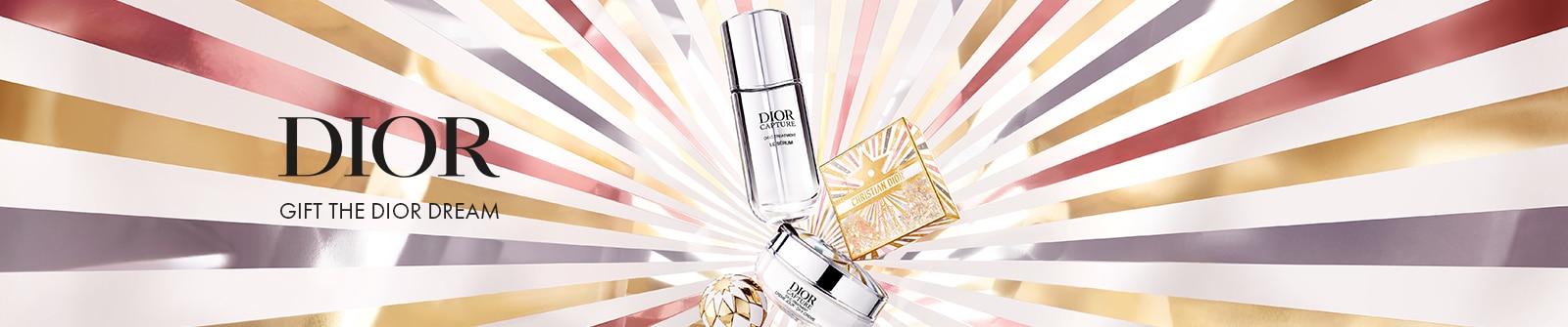 Dior: Gift the Dior Dream - Eine hohe Flasche mit der Aufschrift ‚Capture Totale Le Sérum‘, ein rundes Tiegel mit ‚Capture Totale‘ und eine dekorative quadratische Box vor einem strahlenden Hintergrund aus goldenen, silbernen und bronzenen Sternstrahlen.