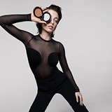 Frau trägt ein eng anliegendes schwarzes Outfit mit transparenten Mesh-Details, posiert vor einem schlichten hellgrauen Hintergrund und hält ein geöffnetes Lancôme Teint Idole Ultra Wear Bronzepuder-Compact vor ihrem Auge.