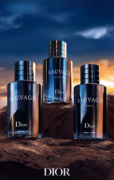 Drei Flaschen des Dior Sauvage Parfüms – Sauvage, Sauvage Parfum und Sauvage Eau de Parfum – sind auf einer felsigen Oberfläche vor einem dramatischen Sonnenuntergangshimmel arrangiert. 