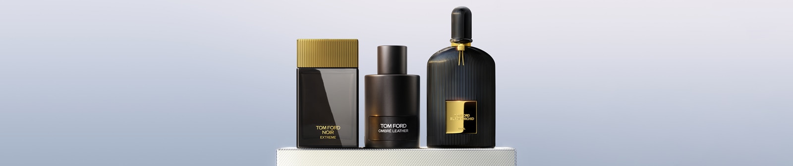 Drei Tom-Ford-Parfumflaschen – Noir Extreme mit goldener Kappe, Ombre Leather in mattem Schwarz und Black Orchid mit geriffeltem Design und goldenen Akzenten – nebeneinander auf einer weißen strukturierten Oberfläche vor einem Farbverlauf-Hintergrund.
