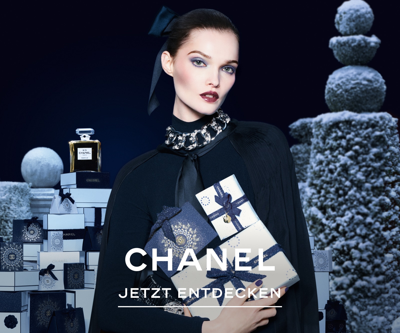 Chanels Winter Constellation-Kampagne mit einem Modell, das drei Chanel-Geschenkboxen hält. Im Hintergrund sind schneebedeckte Formschnittstrukturen und ein Stapel ähnlicher Geschenkboxen zu sehen, gekrönt von einer Flasche Chanel No. 5 Parfum.